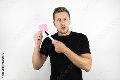 handsome young man office worker wearing black t-shirt holding pink note paper and marker looks confused isolated white background