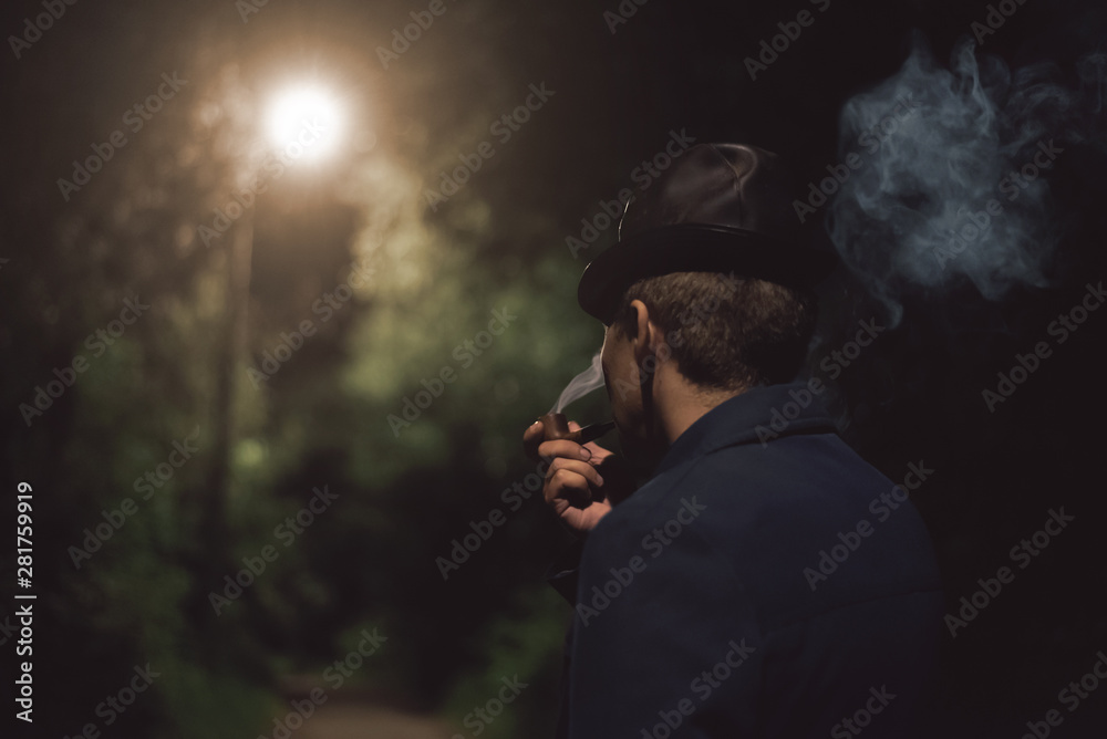 Detective agent in a hat and coat smokes his smoking pipe and thinking in a night park. Detective agent in a hat and coat smokes his smoking pipe and thinking in a night park.