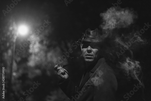 Detective agent in a hat and coat smokes his smoking pipe and thinking in a night park.
