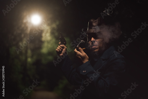 Detective agent in a hat and coat is smokes a smoking pipe with a handgun in a hand.