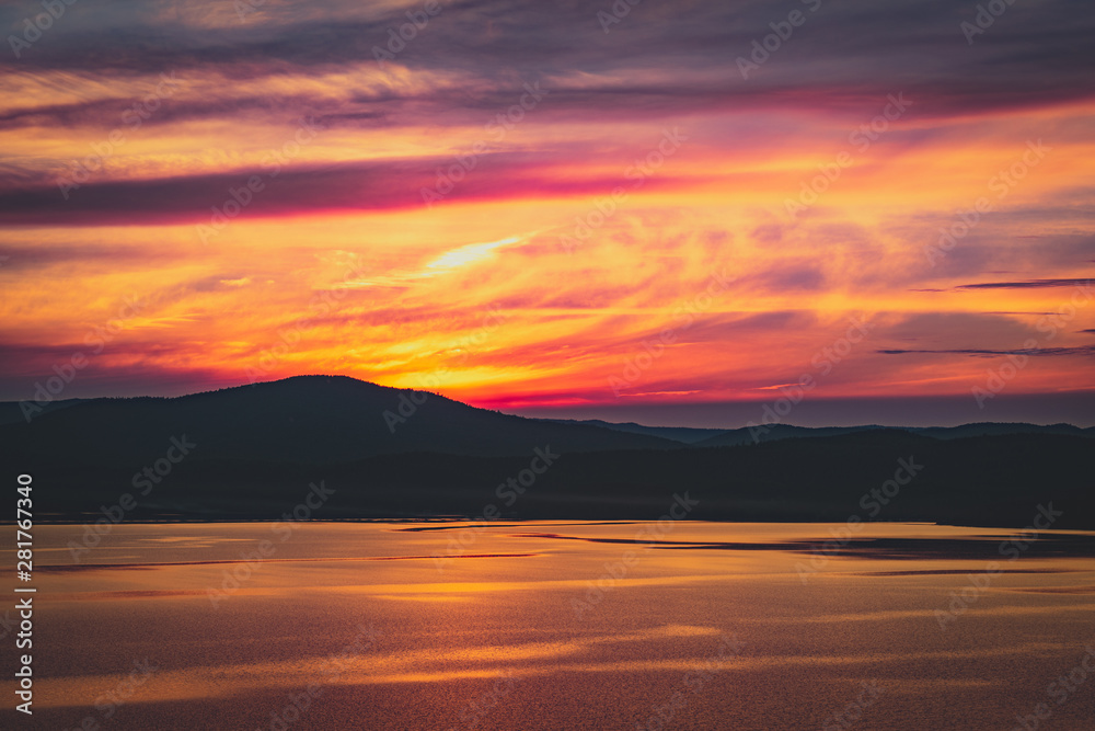 Beautiful burning cloudy sunset at the lake with sun gone behind the mountains