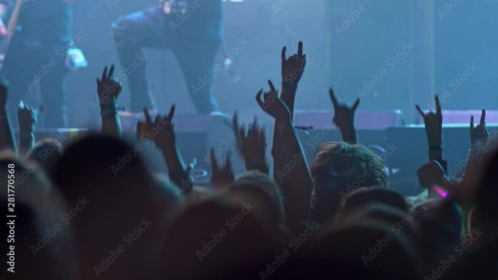 People in a crowd jumping to the groove during a rock concert. They have their hands showing horns gesture. The stage is illuminated with blue light while a band is performing. 4K