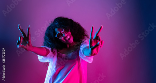 Fashion young african girl black woman wear stylish pink glasses dance look at camera show peace gesture enjoying festive party isolated on disco purple studio background, portrait, copy space