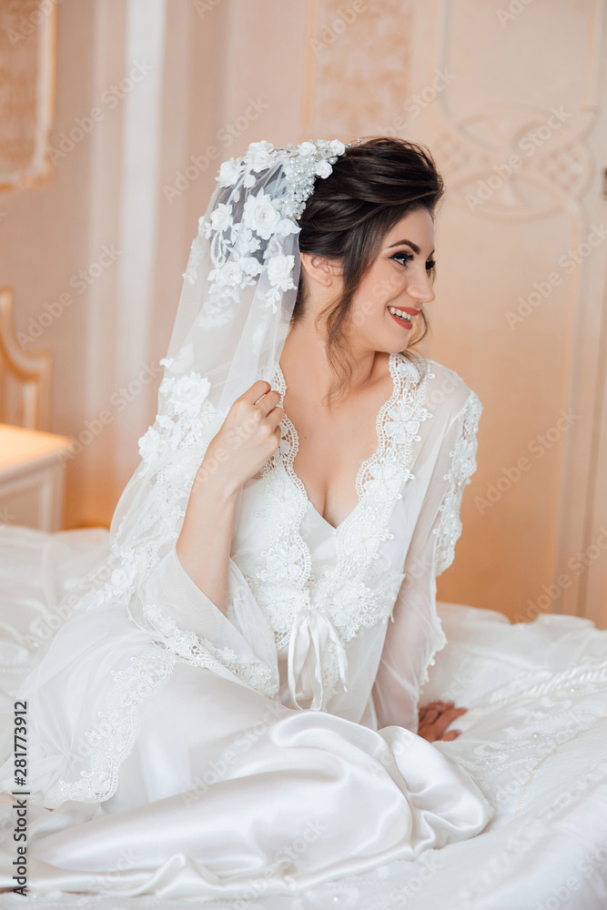 Young luxurious brunette sitting on a bed in a white robe. Morning of beautiful bride. Bride's fees. Elegant hairstyle and makeup. Young luxurious brunette sitting on a bed in a white robe. Morning of beautiful bride. Bride's fees. Elegant hairstyle and makeup.