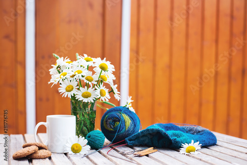 Women's hobby. Crochet and knitting. Workspace. A bouquet of daisies in a vase, a white Cup with a drink, cookies, yarn balls, needles, scissors, knitwear, crochet hooks on a wooden table in a cozy ho