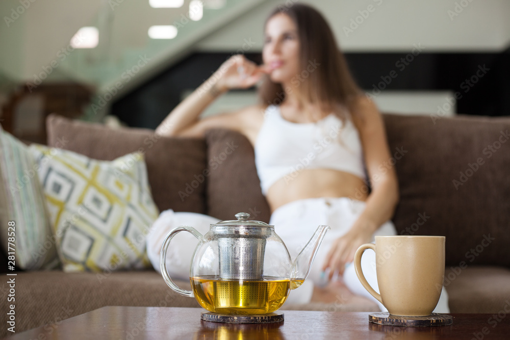 glass teapot with herbal tea and mug on the table. unfocused beautiful smiling girl sitting on the sofa in the background glass teapot with herbal tea and mug on the table. unfocused beautiful smiling girl sitting on the sofa in the background