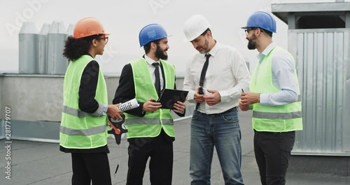 Charismatic young African lady multiethnic group of workers on the top of construction site they chatting with each other analyzing the plan of building wearing the hard hat safety helmets photo