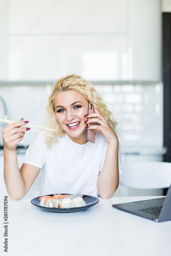 Young blonde woman talking on the phone sitting at her kitchen eat sushi at table home working with a laptop in the morning Young blonde woman talking on the phone sitting at her kitchen eat sushi at table home working with a laptop in the morning