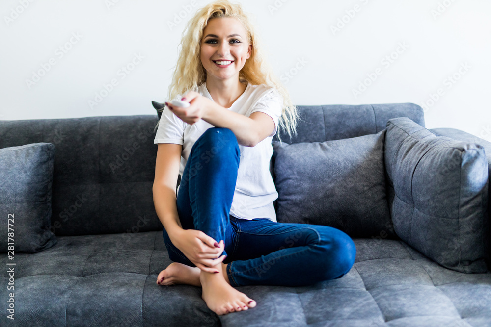 Young smiling woman holding a remote control at home sitting on the couch and watching tv