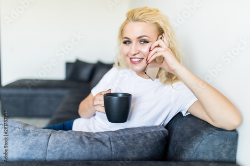 Pleasant talk with friend. Attractive young brunette woman talking on the phone and holding coffee cup while sitting on the sofa. Technology, communication and coziness concept