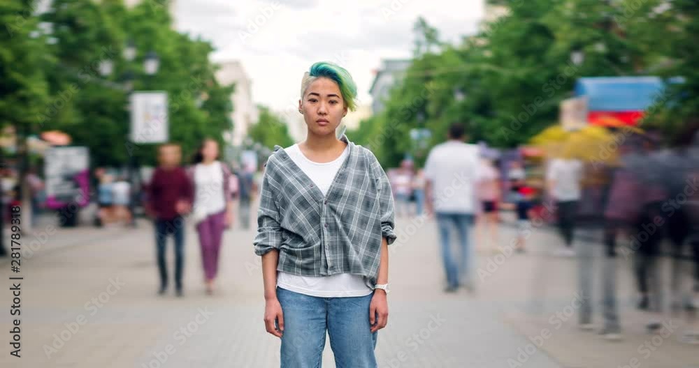 Time lapse of beautiful Asian teen with dyed hair looking at camera outdoors standing alone among flow of pedestrians. Modern lifestyle and loneliness concept. Time lapse of beautiful Asian teen with dyed hair looking at camera outdoors standing alone among flow of pedestrians. Modern lifestyle and loneliness concept.