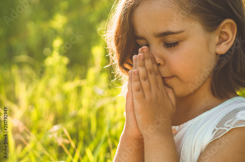 Little Girl closed her eyes, praying in a field during beautiful sunset. Hands folded in prayer concept for faith, spirituality and religion. Peace, hope, dreams concept