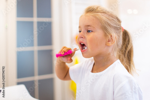 Portrait of cute little girl with blonde hair which cleaning tooth with brush and toothpaste in bathroom.