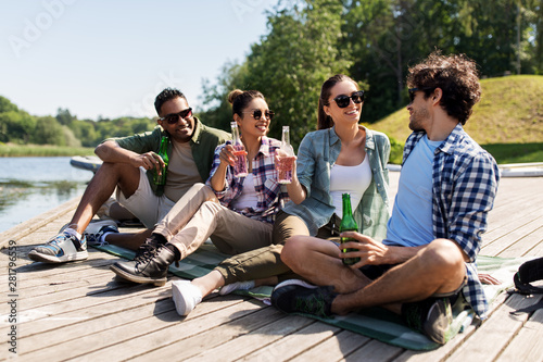 leisure, picnic and people concept - friends drinking beer and cider on lake pier in summer park