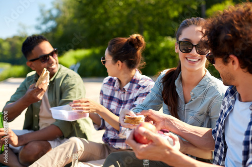 leisure and people concept - group of happy friends having picnic and eating sandwiches on lake pier in summer