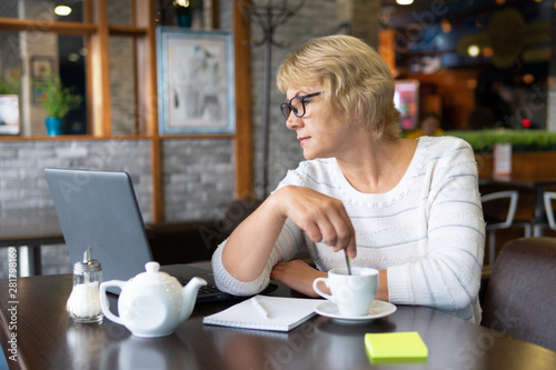 A middle-aged woman with a laptop works in a cafe in the office, she is a freelancer. A woman with glasses sits at a table with a Cup of herbal tea. She is reading.