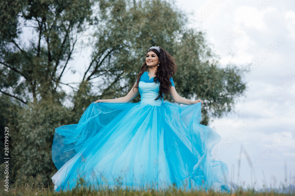 Portrait fashion girl standing near tree. Amazing brown hair model in blue dress looking at camera and posing. Fairytale about young princess.Perfect make up. Young elegant woman in blue long flying Portrait fashion girl standing near tree. Amazing brown hair model in blue dress looking at camera and posing. Fairytale about young princess.Perfect make up. Young elegant woman in blue long flying