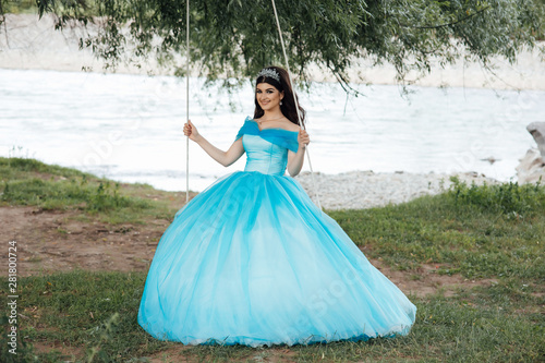 The girl in a blue luxury dress is riding a swing near river. Young carefree girl swinging on a wooden swing and looking at the camera. Perfect make up, healthy wavy hairs. Fashion portrait.