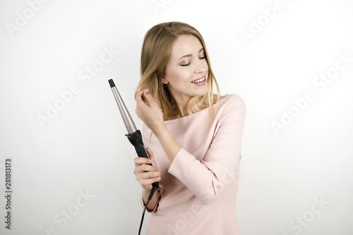 young beautiful blonde woman wearing trendy pink dress using curling iron to make fashionable hairstyle on isolated white background