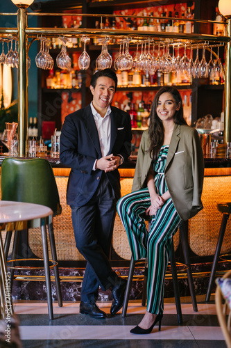 Portrait of two professionally and well-dressed young Asian individuals out on a date at a classy bar together. One is a Chinese man in a suit with his elegant Indian woman date -- they are talking.