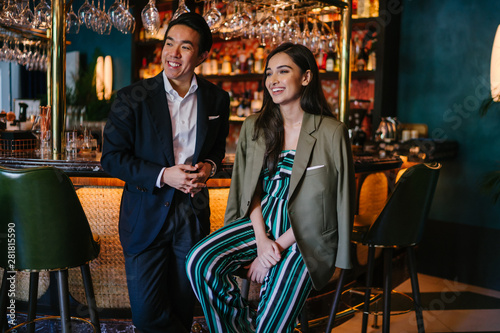 A photogenic young diverse Asian couple enjoy a drink at a classy bar. A Chinese man in a well-tailored and professional suit is talking with an elegant Indian woman who is laughing out loud. 