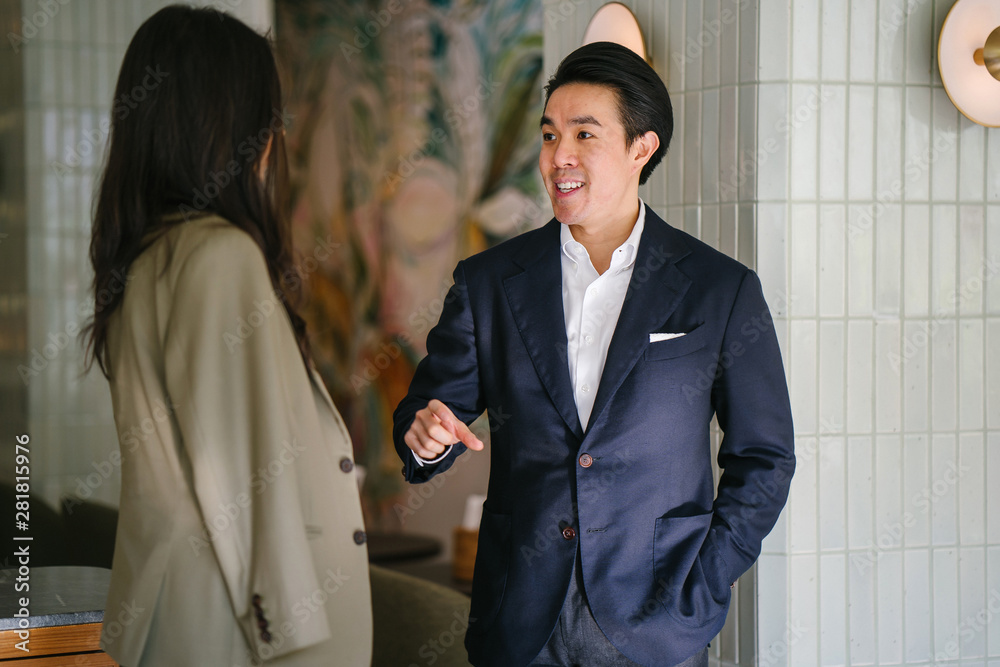 Φωτογραφία A young and attractive Chinese Asian man in a well-fitted navy suit and pocket square is smiling and gesturing as he talks to an Indian Asian woman in a jacket