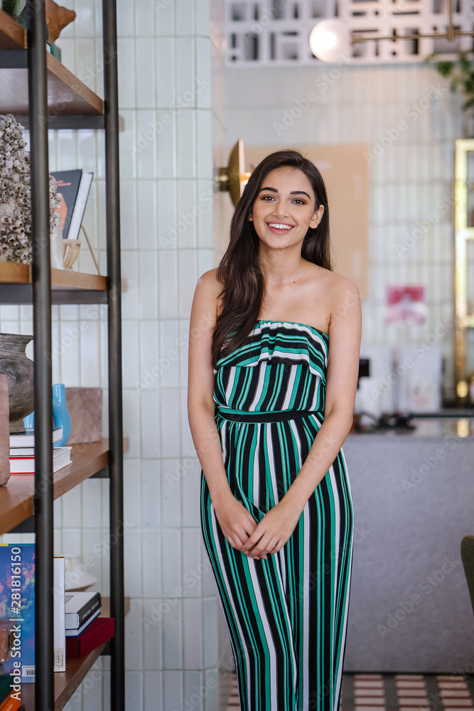 Φωτογραφία Portrait of a beautiful, sophisticated and attractive young Indian Asian woman (high society) in an elegant green outfit smiling as she stands next to a bookshelf in her well-appointed home