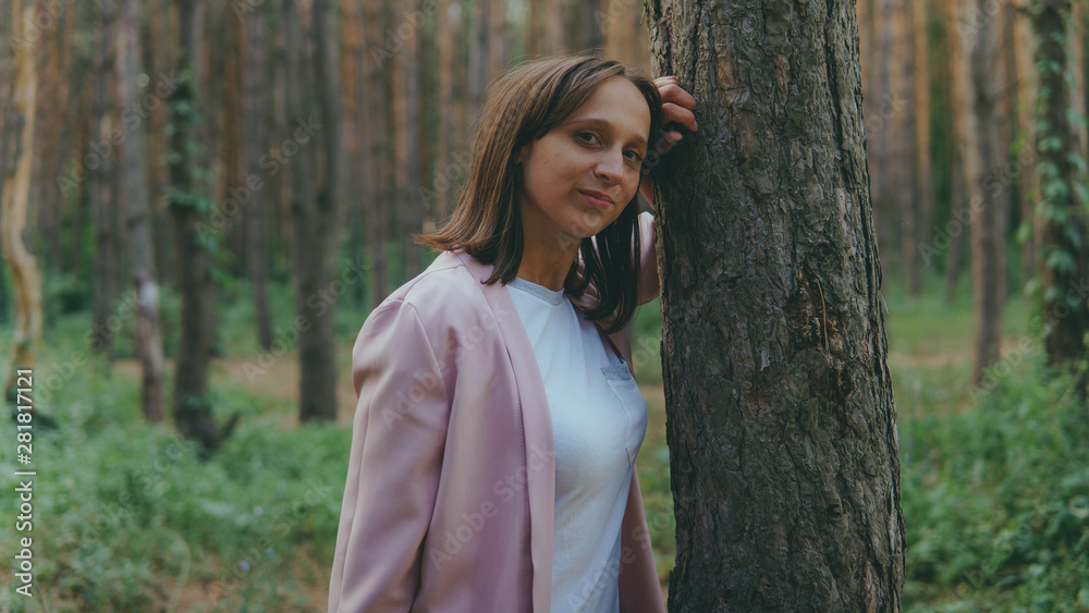 Serious woman looking out from behind tree. Pretty calm woman touching tree and looking at camera in quiet forest Serious woman looking out from behind tree. Pretty calm woman touching tree and looking at camera in quiet forest
