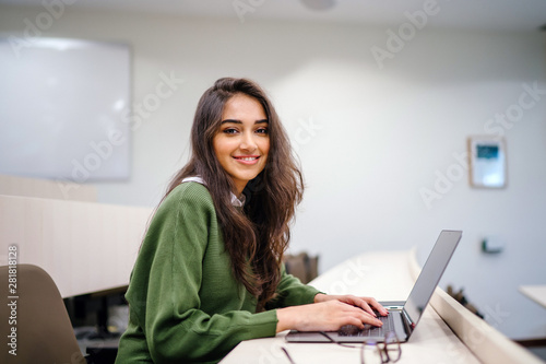 Portrait of a beautiful, young and intelligent-looking Indian Asian woman student wearing a white shirt and green tracker smiling as she works on her laptop in a university classroom.