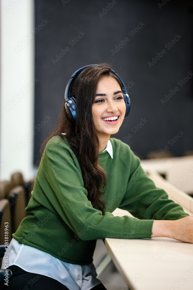 Ταπετσαρία Portrait of a young and attractive Indian Asian woman student in a preppy outfit sitting in a classroom and smiling as she listens to music streaming from her phone to her wireless headphones