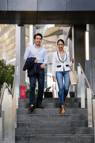 Two young professionals walk side by side. One is a Chinese Asian man in a suit and the other a woman in a white suit -- they are both engaged in conversation as they walk in the city together.