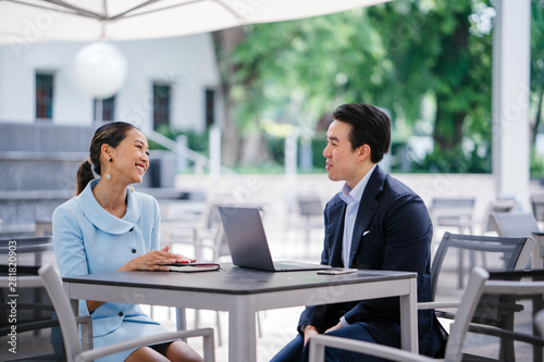 An Asian business man in a crisp suit interviews a candidate (a woman in blue suit) during the day in the city. He is sitting with his laptop in front of him to take notes as he talks to the candidate