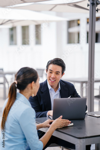 A young, handsome and well-groomed Asian man in a dark suit is interviewing a professional woman candidate applying a job during the day. He is smiling as he talks to the woman.