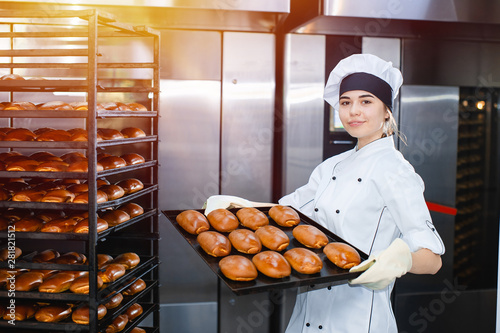 Young girl baker holds a baking tray with hot pastries on the background of an industrial oven in a bakery.