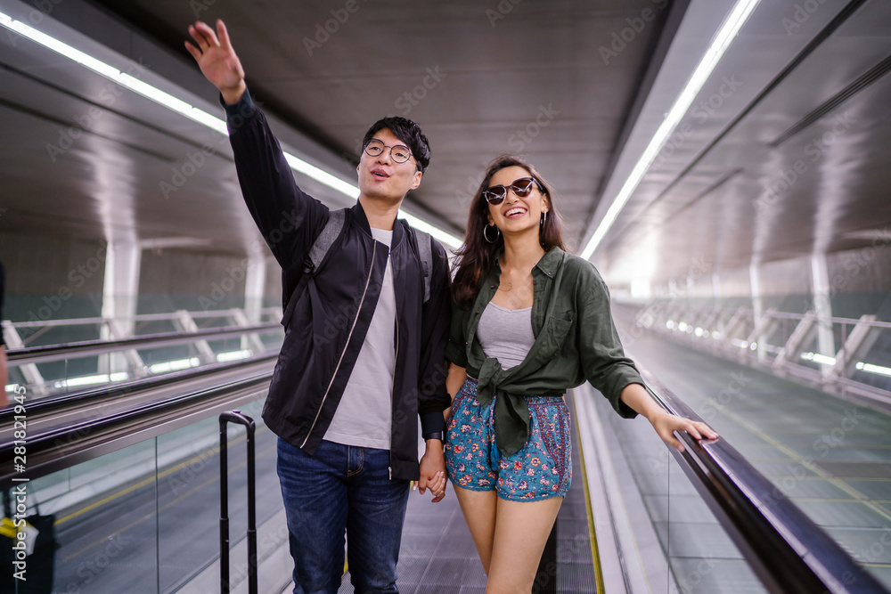 A young and attractive diverse interracial Asian couple (Korean man, Indian woman) are on their way for a holiday. They are in the airport and smiling while they point excitedly.