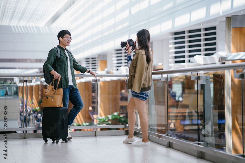 Obraz premium Portrait of a young, diverse and casually dressed interracial Asian couple (a Korean man and Indian woman) standing and taking pictures of one another in a futuristic airport during the day.