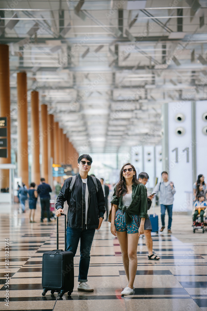 Obraz premium A young and photogenic Asian couple (Korean man, Indian girlfriend in sunglasses) smile as they walk in an airport. They are pulling their luggage behind them on their way for a vacation overseas.