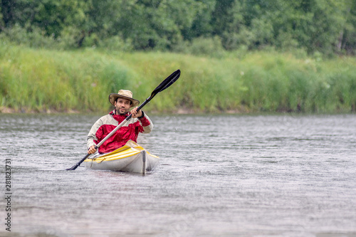 Front view portrait kayaker in round hat on summer river landscape with heavy rain