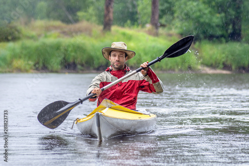 Front view portrait kayaker in round hat on summer river landscape with heavy rain