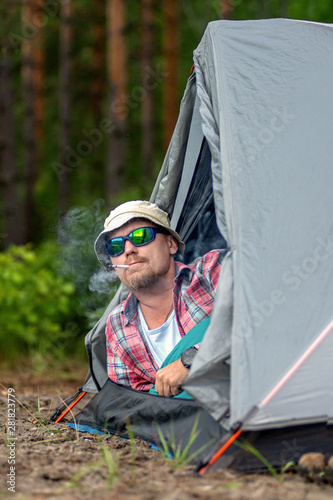 man in a hat and sunglasses lying in a tent and smoking a cigarette. concept of travel and camping