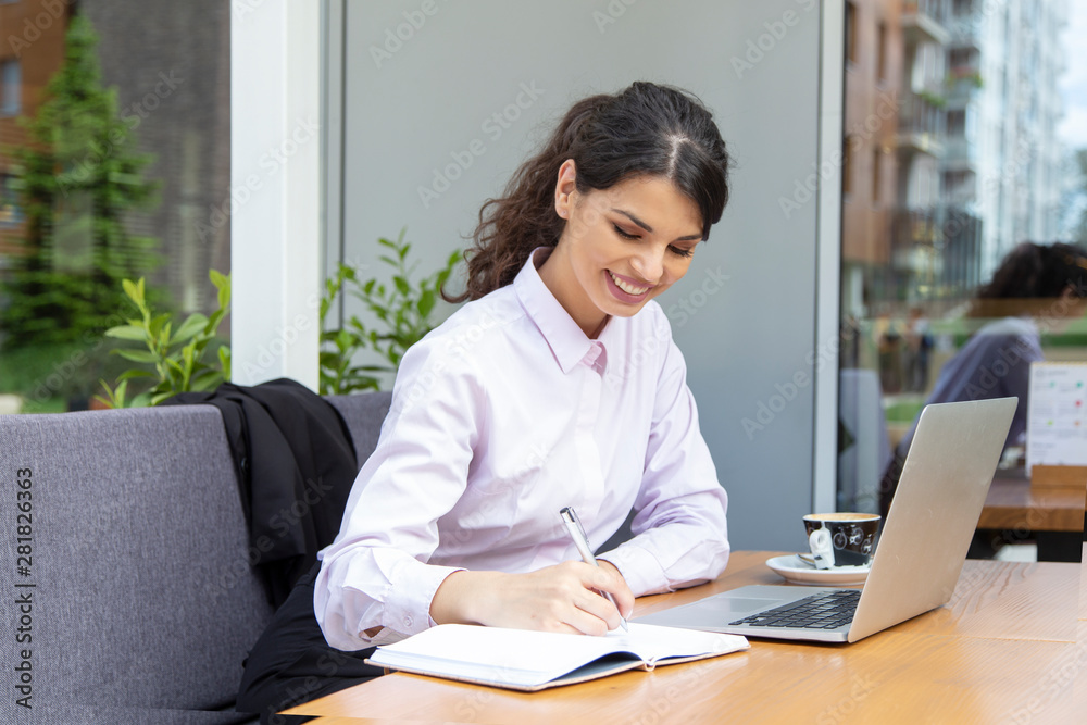 Beautiful young woman work in caffe. Woman writing and using laptop while sitting in restaurant Beautiful young woman work in caffe. Woman writing and using laptop while sitting in restaurant