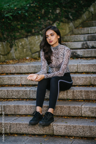 A young Indian Asian woman is sitting on some steps in the park during the evening. She is beautiful, attractive, athletic and tall and is wearing fitting clothes including a snakeskin top.