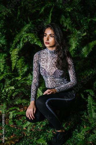 Fashion portrait of a young, beautiful and elegant Indian Asian woman in a snakeskin top and black pants crouching between lush green plants in the night. photo