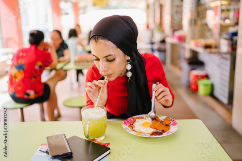 A young Muslim woman in a hijab head scarf eats her halal nasi lemak lunch during the day at a table in a hawker center.