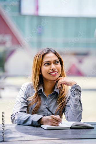 Portrait of a tanned and professional young Southeast Asian business woman in a collared shirt sitting on a bench in the city. She is smiling as she writes and jots in her notebook. Free Download Portrait of a tanned and professional young Southeast Asian business woman in a collared shirt sitting on a bench in the city. She is smiling as she writes and jots in her notebook. - GFXLoad