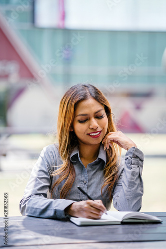 Portrait of a tanned and professional young Southeast Asian business woman in a collared shirt sitting on a bench in the city. She is smiling as she writes and jots in her notebook.