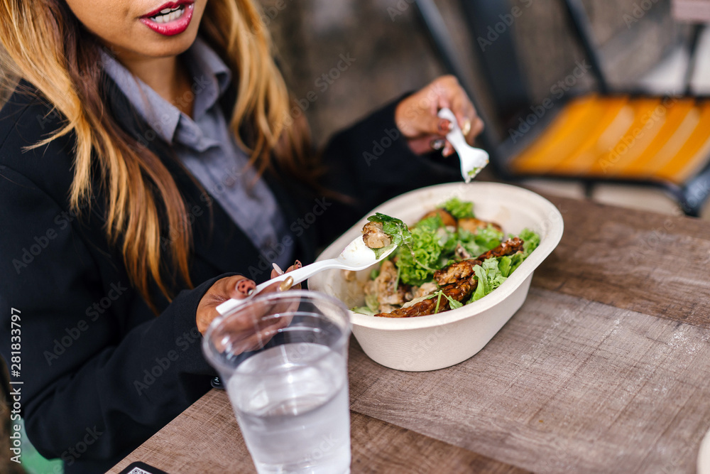 Close up of a young and a attractive Asian woman in a suit eating a healthy salad lunch with a fork and spoon from a takeaway box. She has a glass of water to go with her salad.