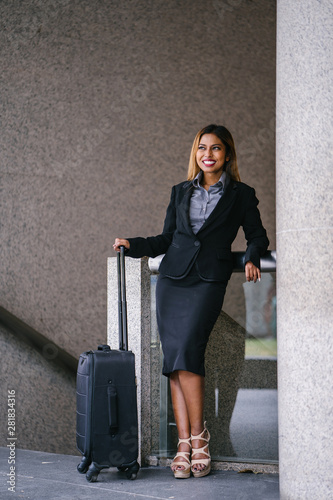 Portrait of a young, beautiful and tanned business woman in a suit smiling confidently as she stands in the city during the day. She is standing next to her luggage to go on a business trip.
