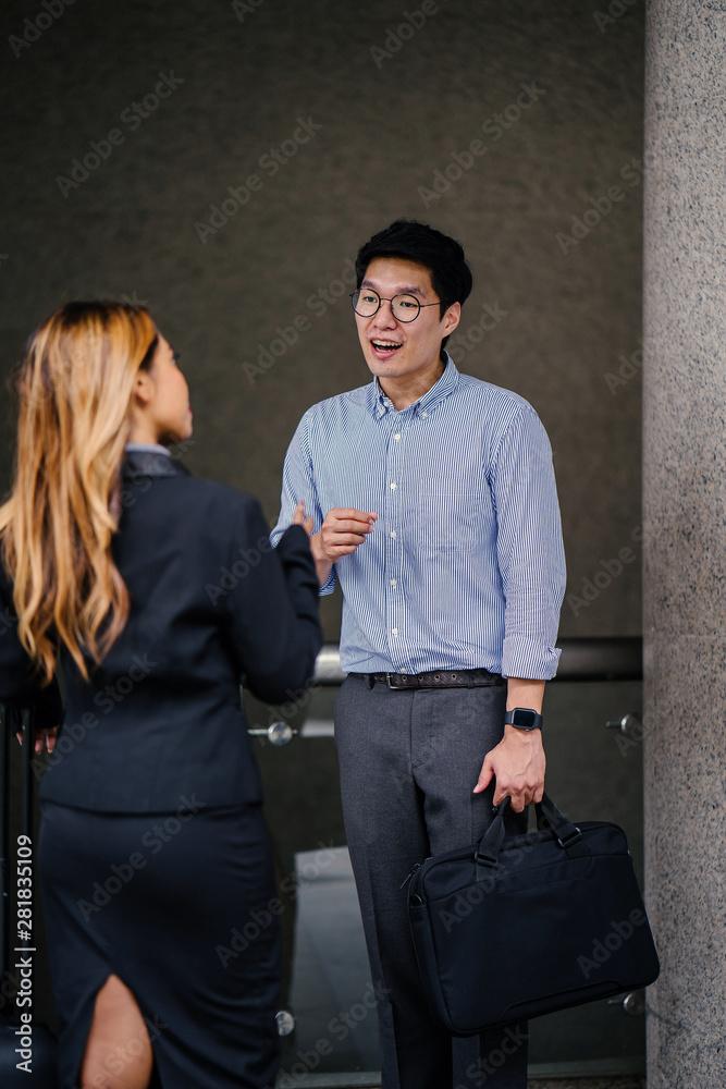 A tall, handsome young Korean Asian man in business attire is talking to his colleague or client (a woman in a business suit) in the city in the day. He is smiling as he speaks to his associate. A tall, handsome young Korean Asian man in business attire is talking to his colleague or client (a woman in a business suit) in the city in the day. He is smiling as he speaks to his associate.