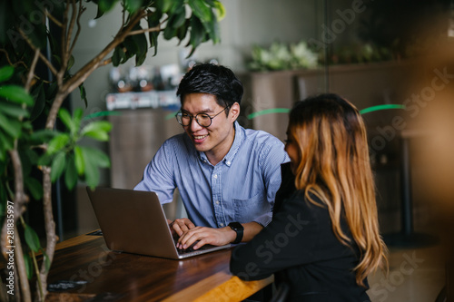 A pair of diverse team mates have a business meeting and discussion together to collaborate. A Korean man is sitting at a table in a trendy coworking space office and talking to his female companion.